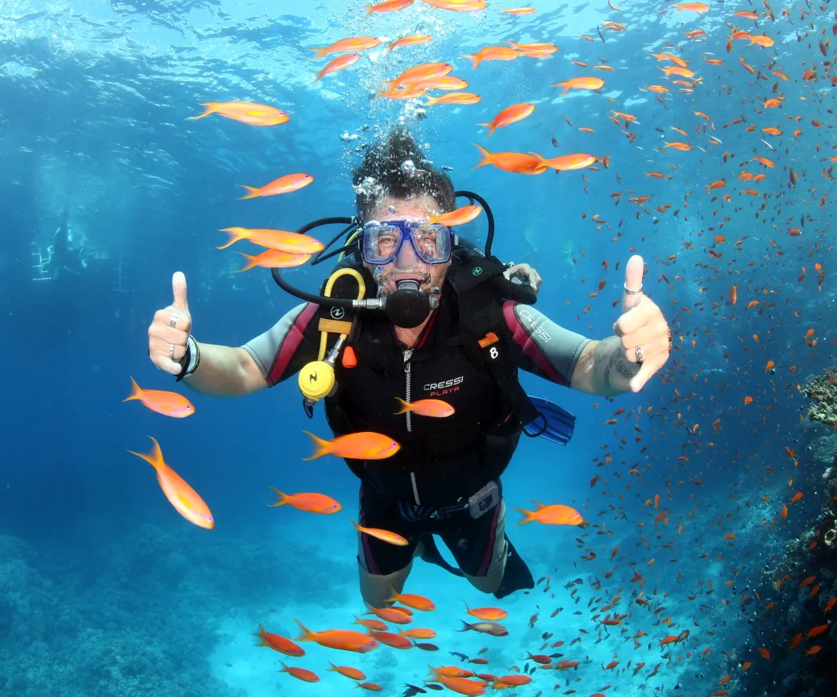 A first-time diver giving two thumbs up among orange anthias on a Red Sea reef