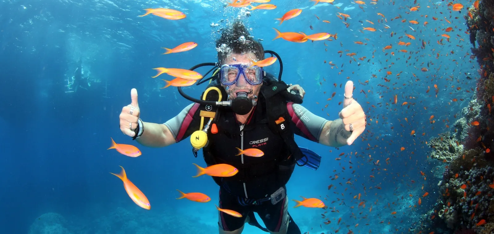 A first-time diver giving two thumbs up among orange anthias on a Red Sea reef