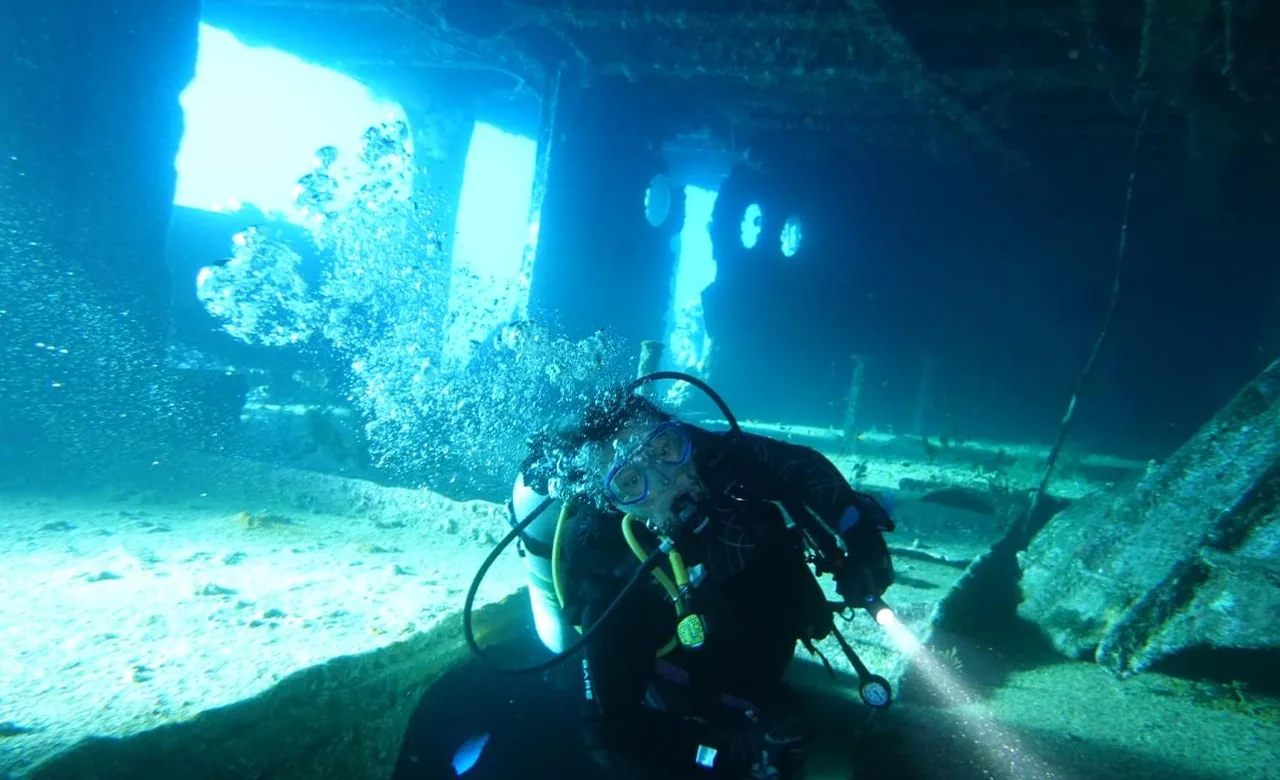 A diver with a torch descending alongside the marine-growth-encrusted hull of an Abu Nuhas wreck